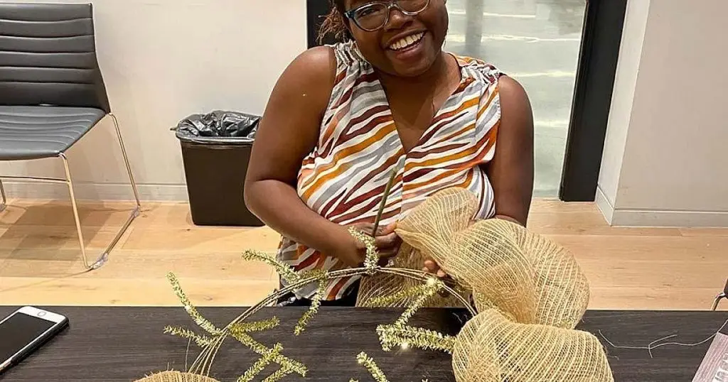 Smiling woman crafting a decorative wreath with mesh ribbon and greenery at a table