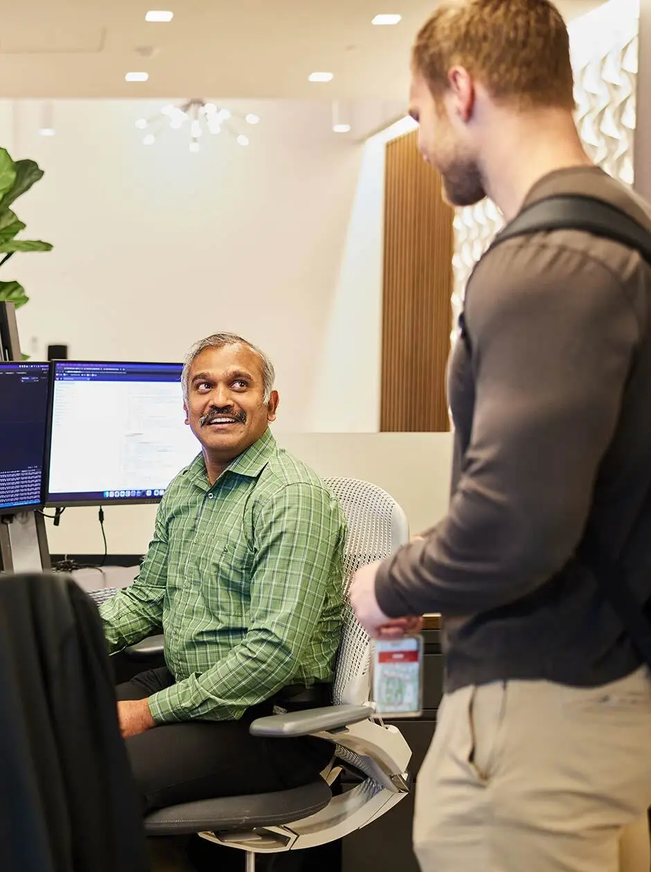 Seated employee chatting with a colleague carrying a backpack in the office